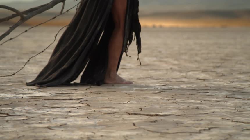 A woman with a cloak dragging a branch in the desert