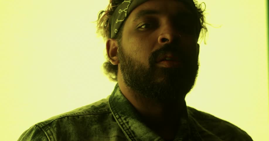 A man with a bandanna around his head and jacket in a room with a big screen in the background