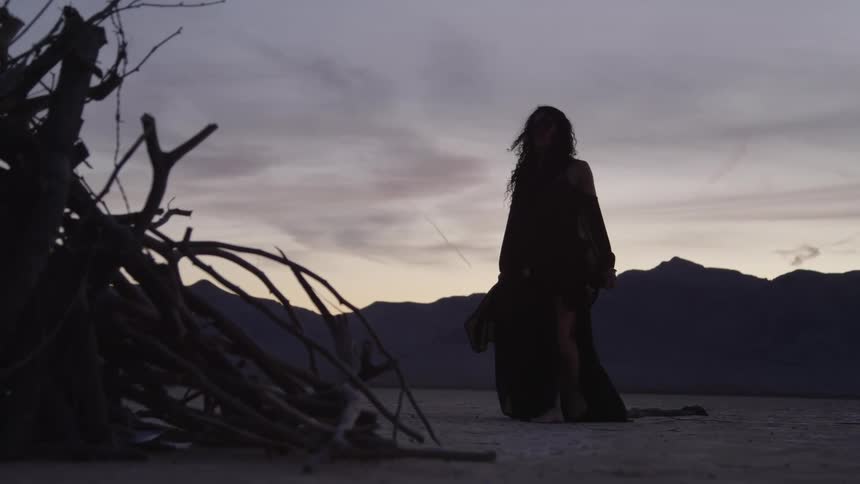 Silhouette of a woman wearing a long dress walking next to a pile of wood in the desert at evening
