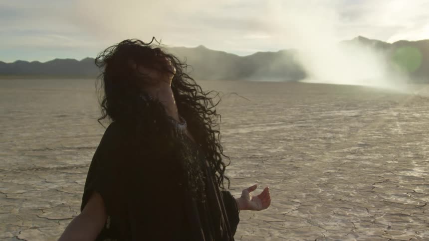 A woman kneeling in the desert with a mountain range the sky and clouds in the background