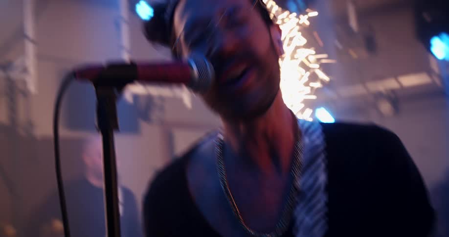 A man sings and plays the guitar on stage with a band as sparks fly into the air behind him