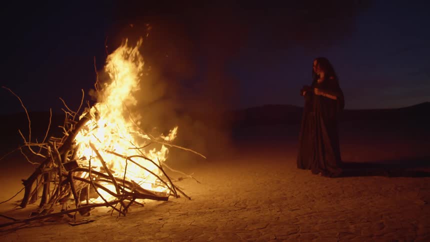 A woman dressed in black with veils stands watching a large fire in the desert