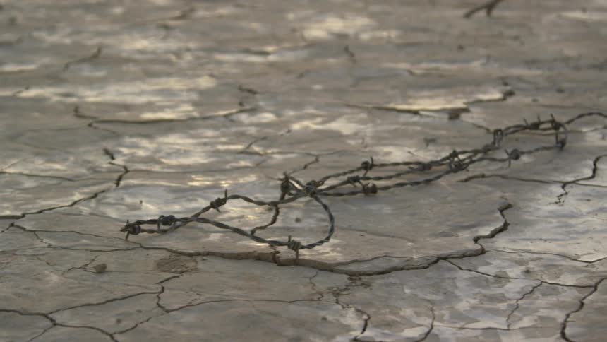 A person in a long black gown drags a branch and barbed wire through the desert