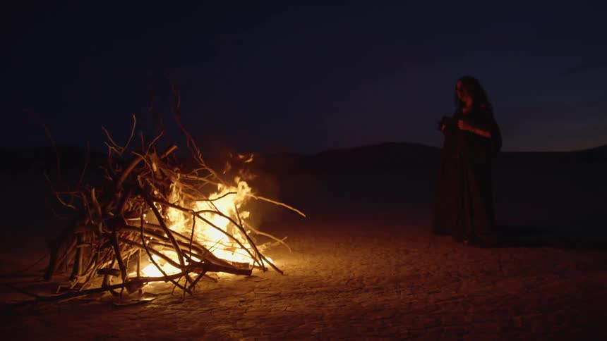 A person is standing near a bonfire watching the flames in the darkness