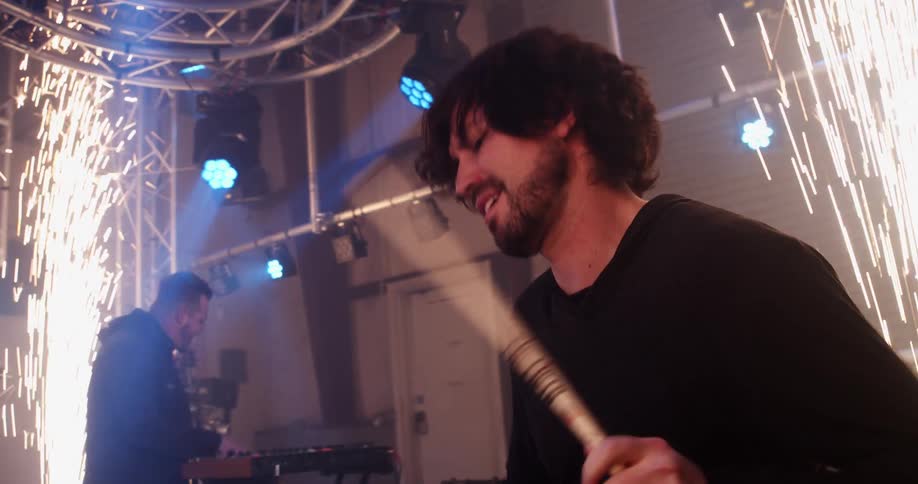 A man on a stage playing drums with drumsticks with another man with a saxophone playing the keyboard in the background
