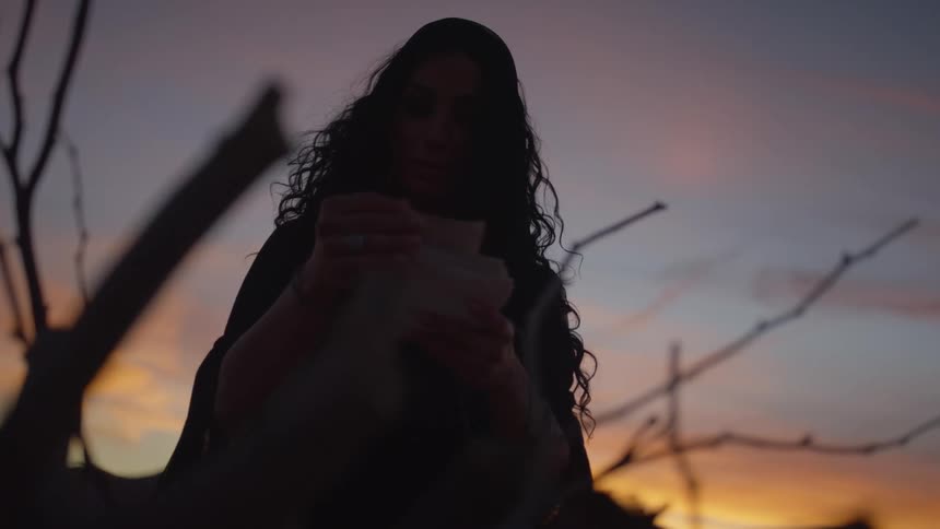 A woman standing in the desert putting pieces of paper by a branch with the sky and clouds in the background