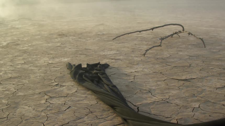 A long black cloth is dragged through arid cracked soil of a flatland through clouds of dust