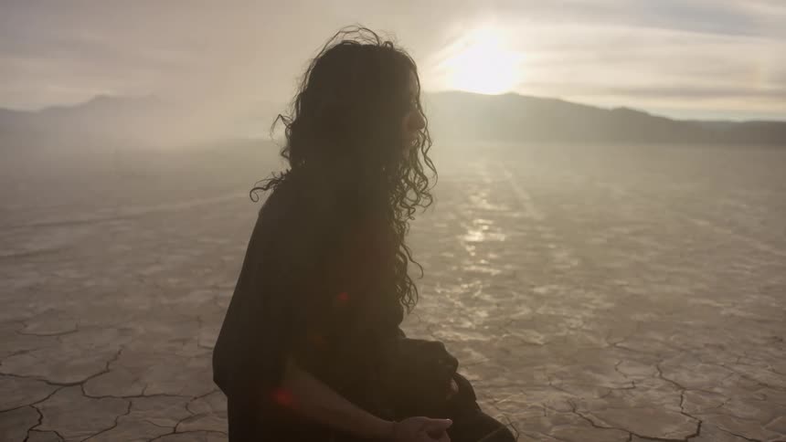 Sunlight shines over a distant mountain onto a woman sitting on the cracked soil of an arid flatland as the wind blows dust around her