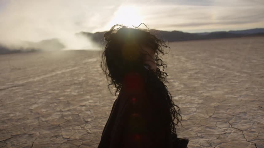 A woman with dark curly hair stands in the desert with a canyon in the distance
