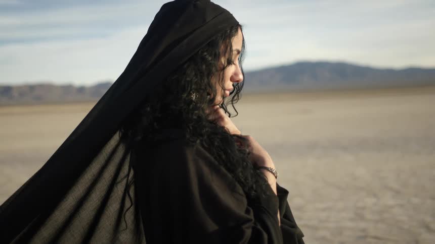 A woman with a dress and veil in a desert with the sky and clouds in the background