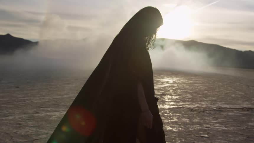 Woman wearing a hooded cloak is walking with a bright sun and mountains in the distance