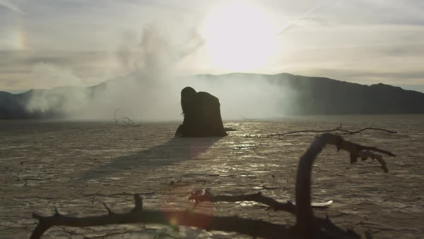 A woman in a long black gown kneels down in the desert and stands back up as smoke fills the air around her