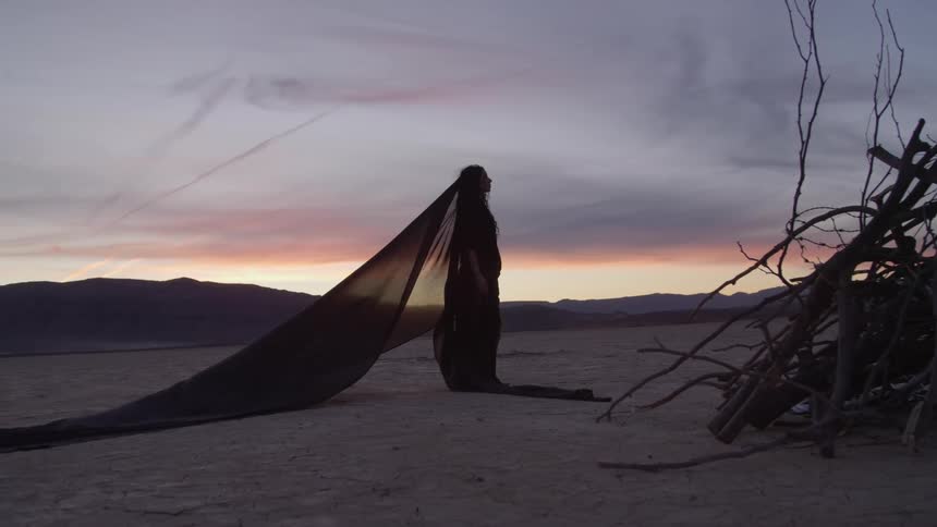 A woman with a long black veil standing alone in a sandy desert at sunrise