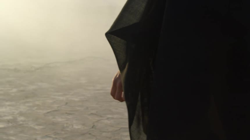 Person wearing voluminous black outfit walks across arid ground toward haze