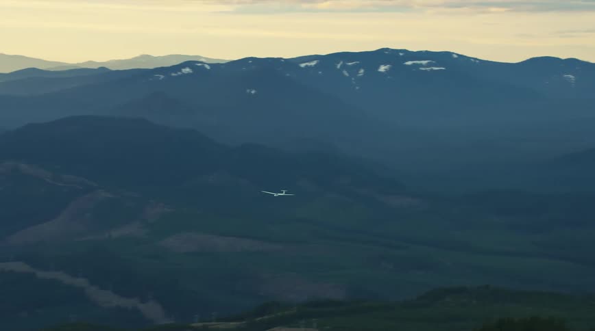 A glider airplane soars high above rugged valleys, ridges and massive mountains with golden sunset light in this air to air aerial