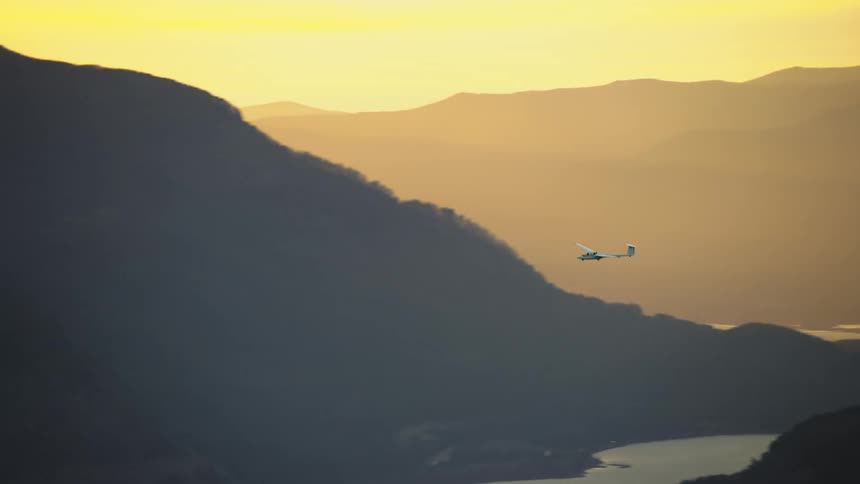 A white glider airplane with no engine soars and glides above a golden hour river gorge and many hills and mountains in this golden hour air to air aerial shot