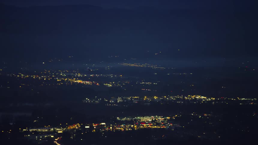 Night air to air shot of a private jet arriving in a large city before landing at a commercial airport under city lights in this blue hour air to air aerial
