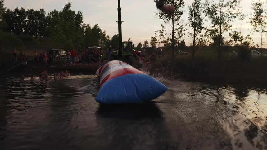 A man laying on a flotation device and being catapulted into the air and falling into a lake with trees the sky and clouds in the background