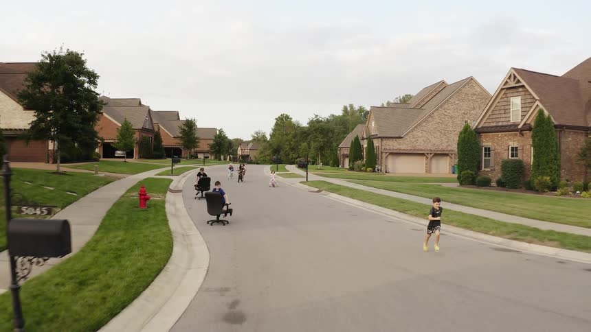 Children running on a neighborhood street with office chairs and bikes with the sky and clouds in the background
