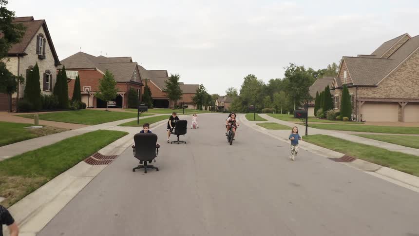 A group of children race down the neighborhood street two pushing desk chairs and one riding a bike as the others run