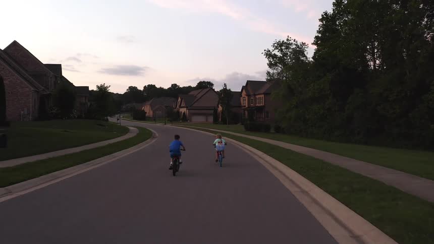 A girl pedals her bike and catches up to a boy riding on a neighborhood road