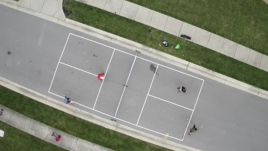 Four people play paddle tennis on a court while kids ride their bikes nearby
