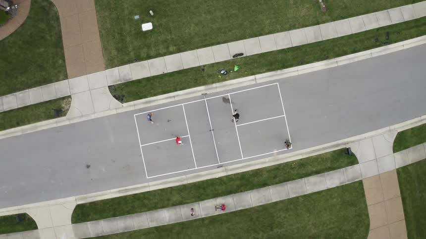 Four people play pickleball ball on a court while a person rides a hoverboard on the walkway