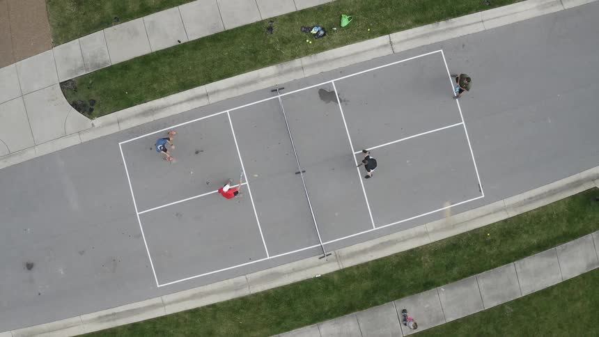 Four people play paddle tennis ball on an outdoor court while a person on a hoverboard rides by