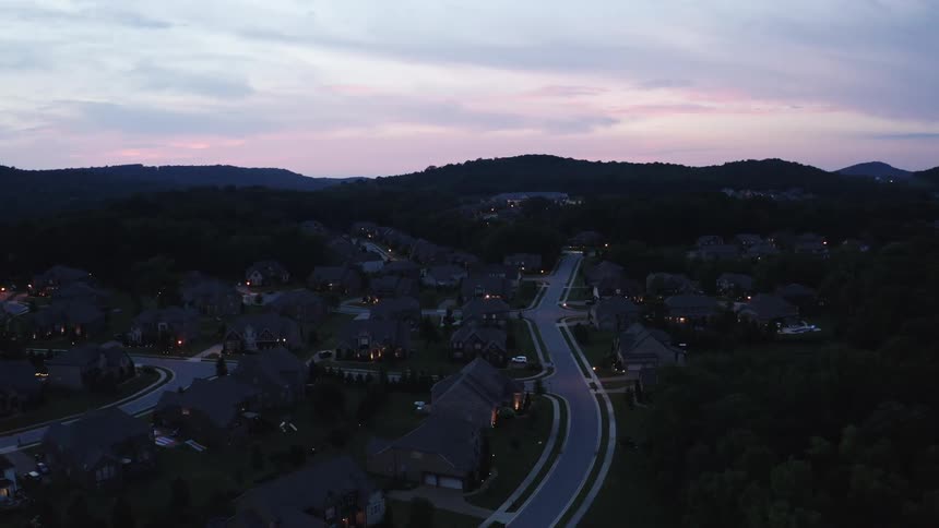 House lights twinkle in a suburban neighborhood with curving road and trees with hills on the horizon