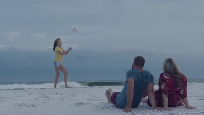Father and mother watching their teenage daugter playing with a volleyball on the white sand of a beach near the ocean