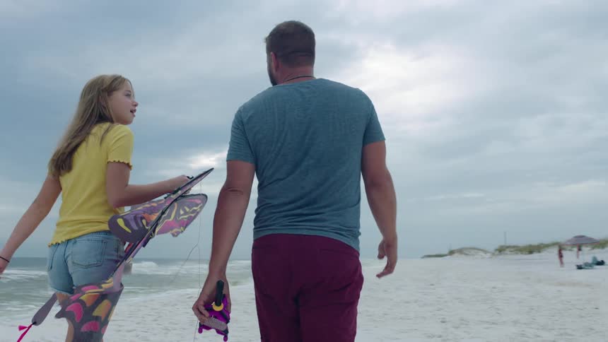 Father and teenage daughter walking on the sand of a beach carrying a butterfly kite