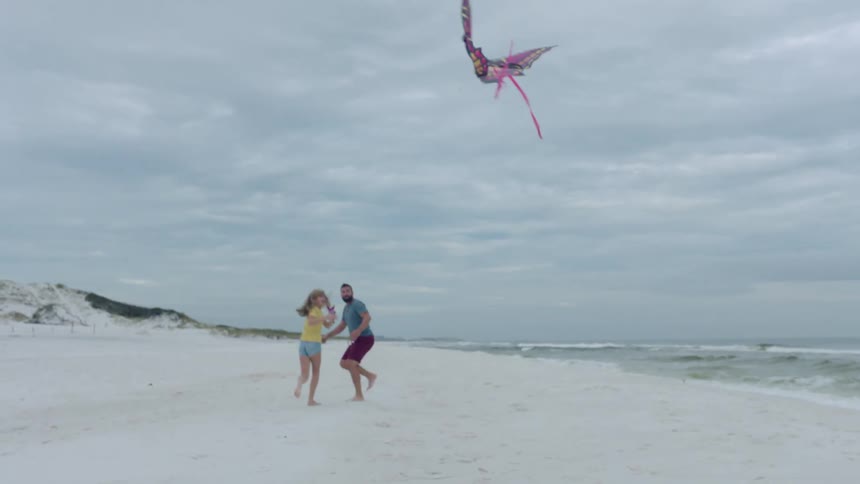 A man runs next to his daughter as she flies a butterfly kite on the beach