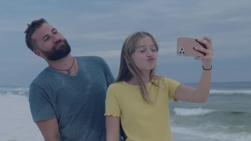 A man and a woman on a beach taking a picture with a smart phone with the sky and clouds in the background