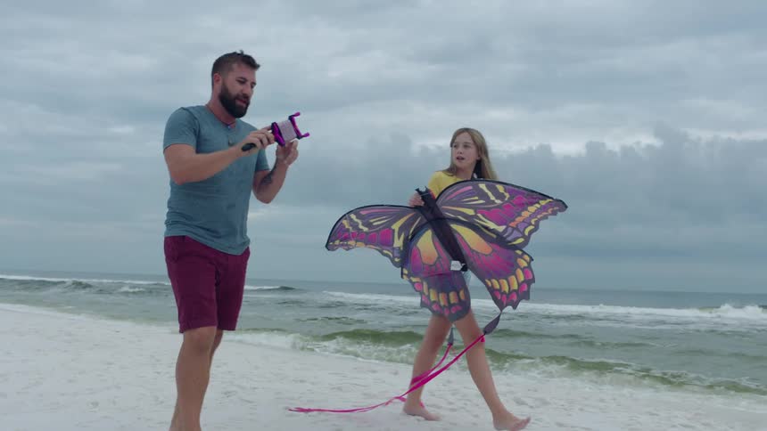 Father and daughter walking with a large butterfly kite along shoreline at the beach