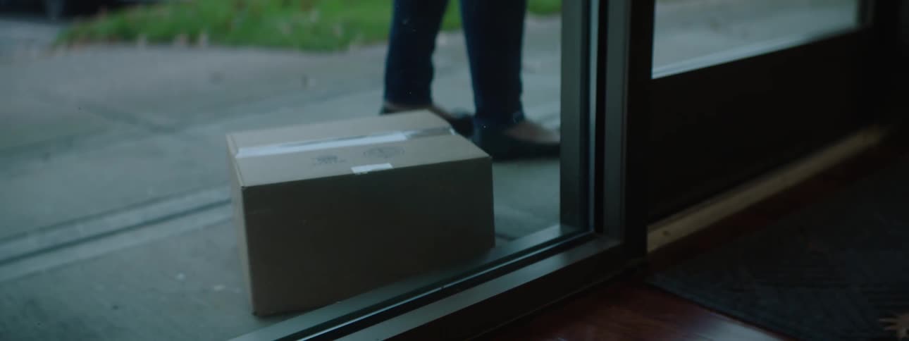 A person placing a box in front of a door with grass and a vehicle in the background