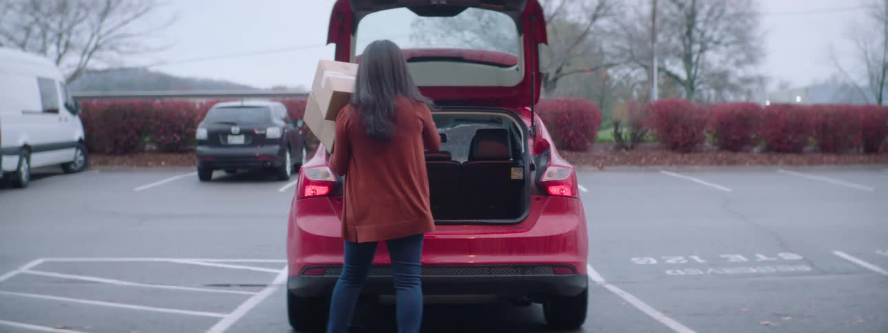 A woman carries a box out to her car and opens the trunk and puts it in