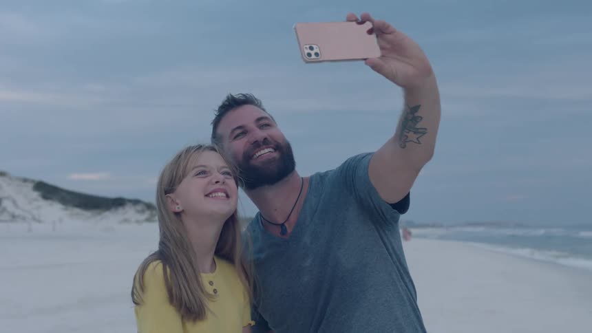 A dad holds up a phone and takes a selfie with his daughter at the beach