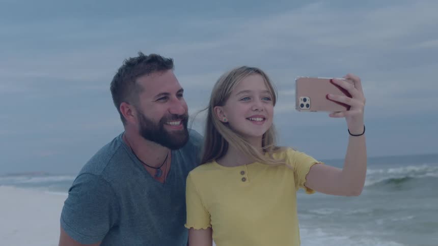 A man and a woman on a beach taking a picture with a smart phone with the sky and clouds in the background