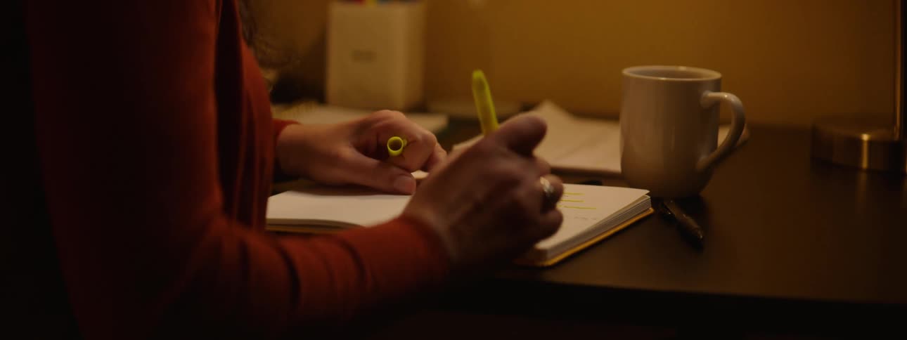 A woman taps her chin with a highlighter as she thinks then highlights lines in her notebook