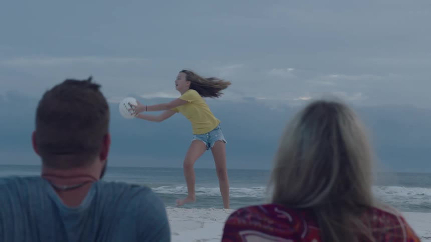 A woman on a beach playing with a ball with a man and a woman sitting in the foreground with the sky and clouds in the background