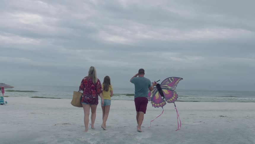 A dad carries a large butterfly kite while walking with his daughter and wife on the beach