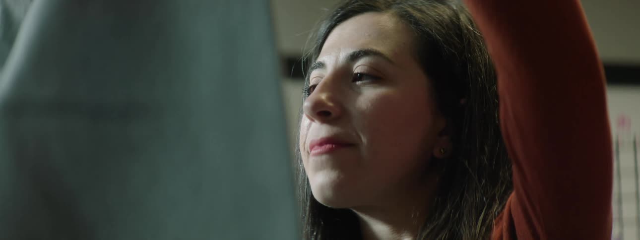 A woman examines a piece of clothing she is holding and smiles in an office with a whiteboard behind her