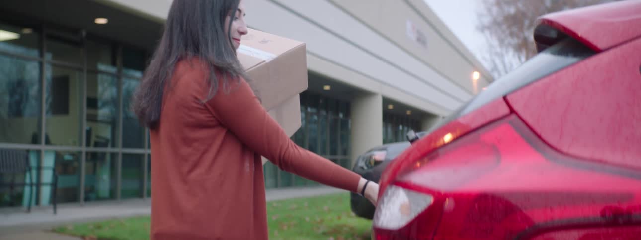 A woman carries two cardboard boxes and loads them into the back of her car