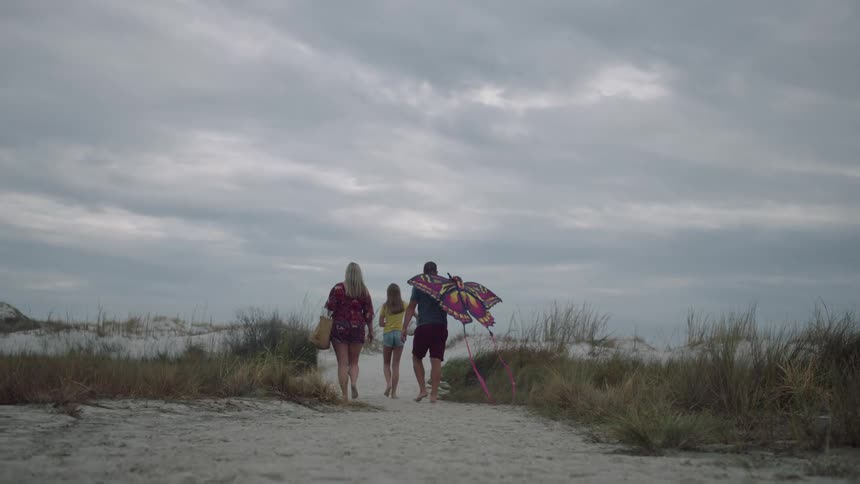 A dad carries a large butterfly kite as he walks on the beach sand with his daughter and wife