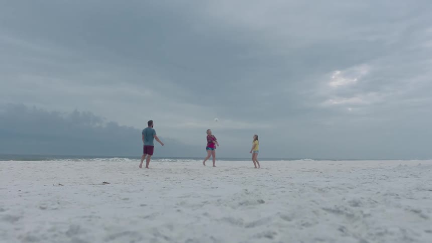A man and two women playing with a ball on the beach with the sky and clouds in the background