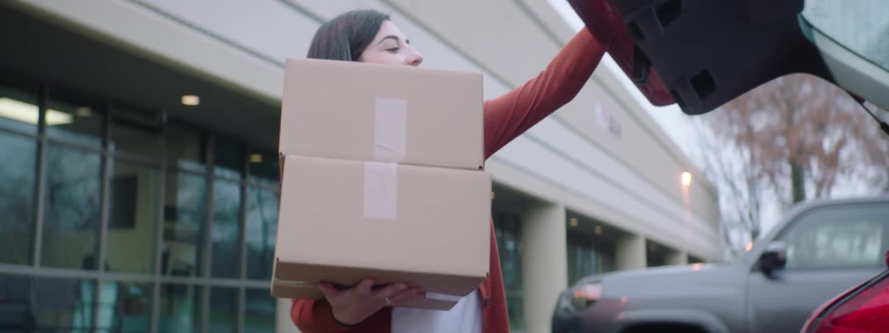 A woman smiles as sheloads two cardboard boxes into her car and shuts the hatchback