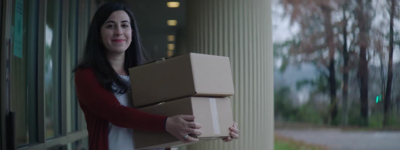 A woman stands outside of a building holding two packages as she smiles