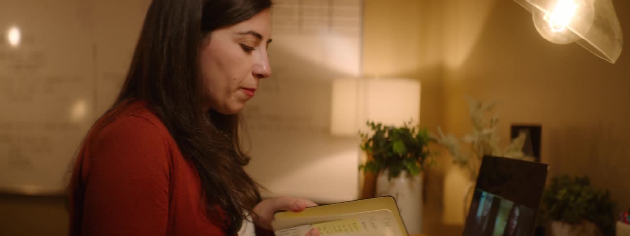 A woman at a desk with a laptop holding a book with a whiteboard in the background
