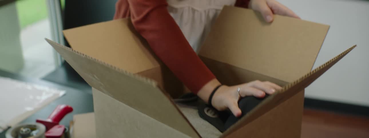 A woman at home packs clothing and a hat in to a box