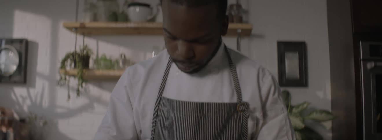 A man wearing an apron and chef's shirt slices cucumbers in a kitchen
